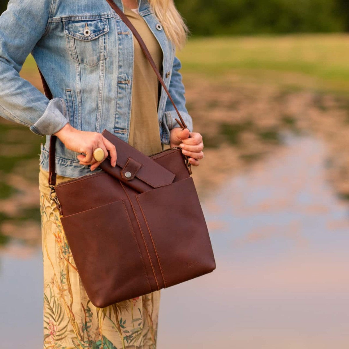 Minimal Leather Bag in reddish brown, worn crossbody by a woman in a denim jacket beside a pond