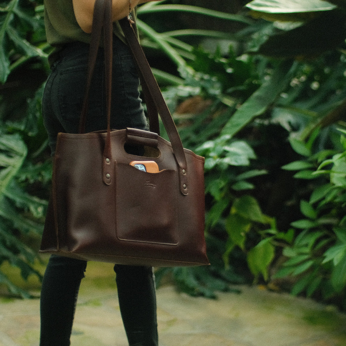 Urban Leather Tote in Chestnut carried on the shoulder by a woman walking past tropical plants