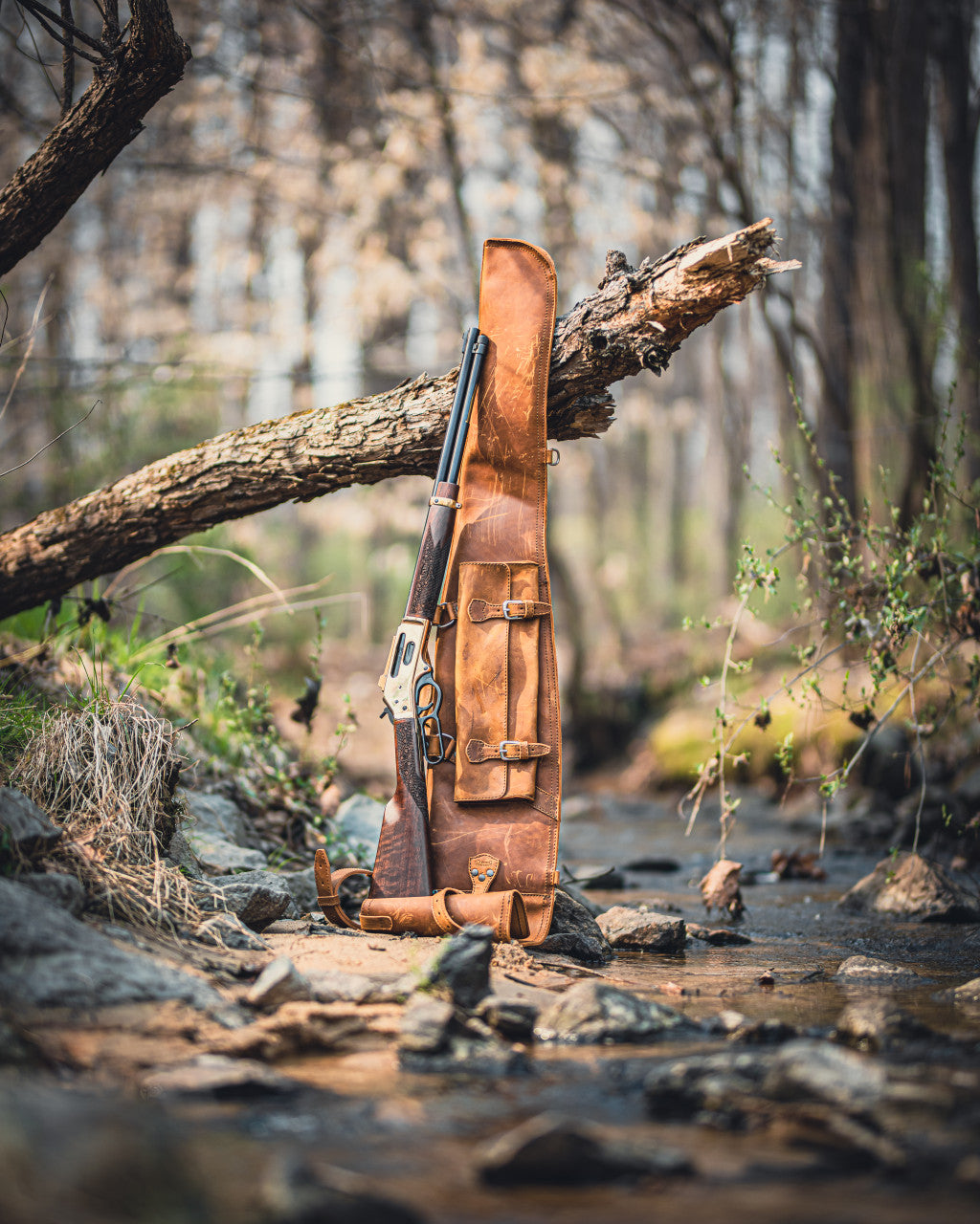 Shotgun Sleeve in Tobacco standing upright by a forest creek beside a lever-action rifle