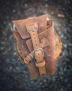 Leather Pistol Wrap in Tobacco folded closed and standing on a tree stump, showing its buckle straps
