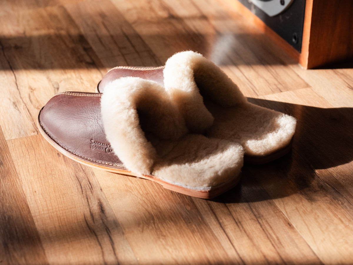 Men's Sheepskin Slippers in brown leather with thick sheepskin lining, in sunlight on a wood floor