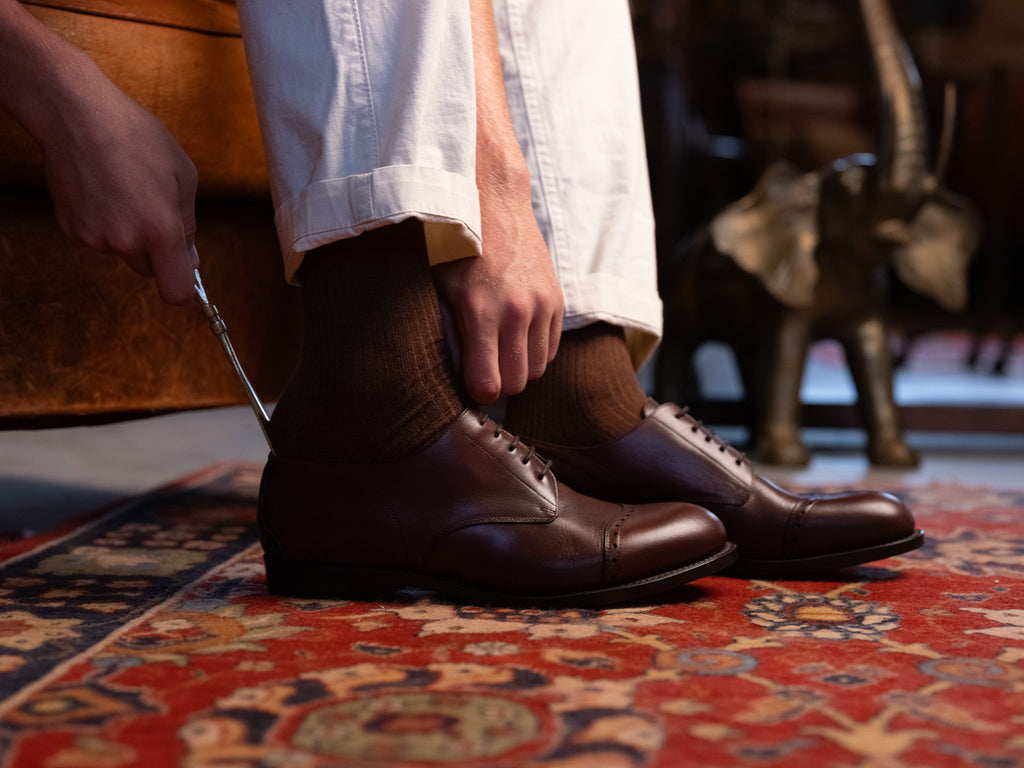 Toe Cap Shoes in Dark Oak, putting them on with a shoehorn on a red patterned rug