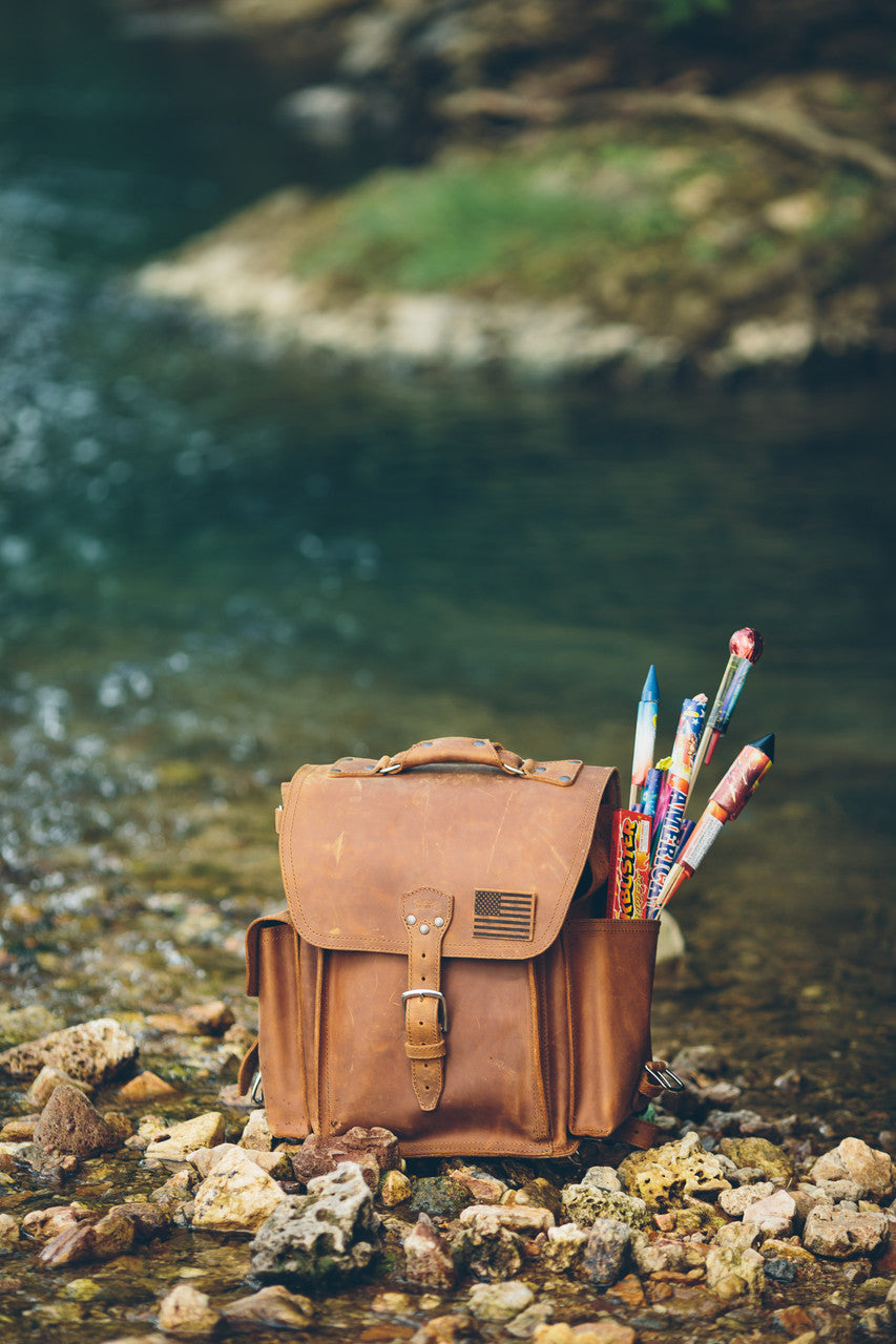 Tank Backpack in Tobacco with an American flag patch, filled with fireworks on a rocky stream bank