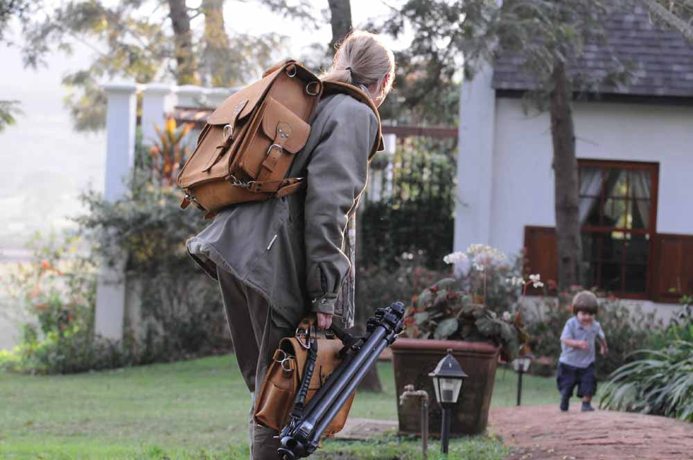 Tank Backpack in Tobacco, carried by a woman with a tripod as a child runs across a garden