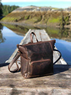 Universal Leather Backpack in Dark Coffee Brown, vertical angled view on a wooden dock by a lake