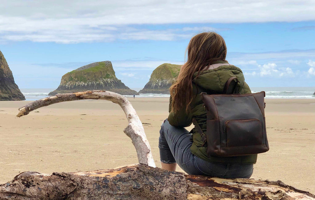Universal Leather Backpack in Dark Coffee Brown worn by a woman sitting on driftwood facing the ocean