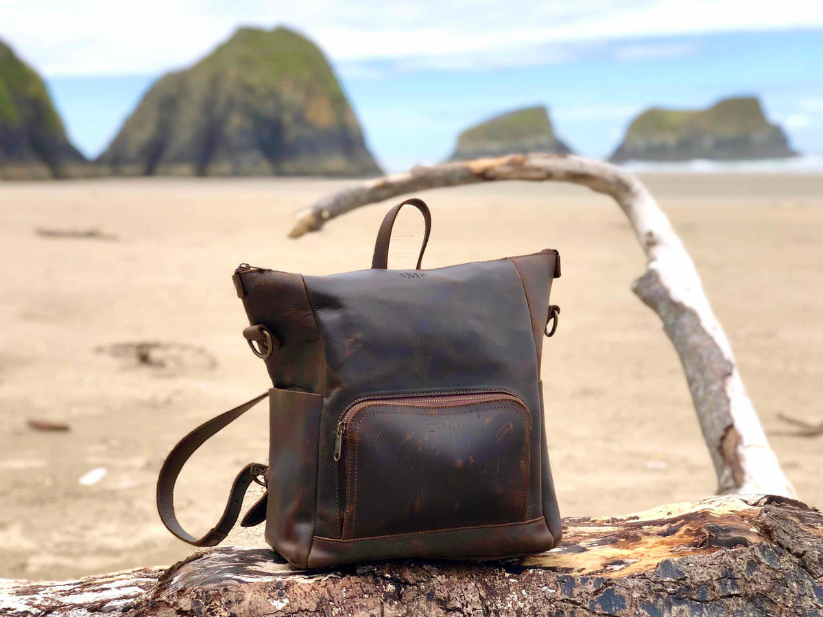 Universal Leather Backpack in Dark Coffee Brown on driftwood at a beach with sea stacks behind