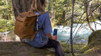 All in One Leather Backpack in Tobacco worn by a hiker sitting on a log beside a forest river