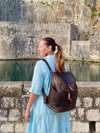 All in One Leather Backpack in Dark Coffee Brown worn by a woman in a blue dress beside old stone walls