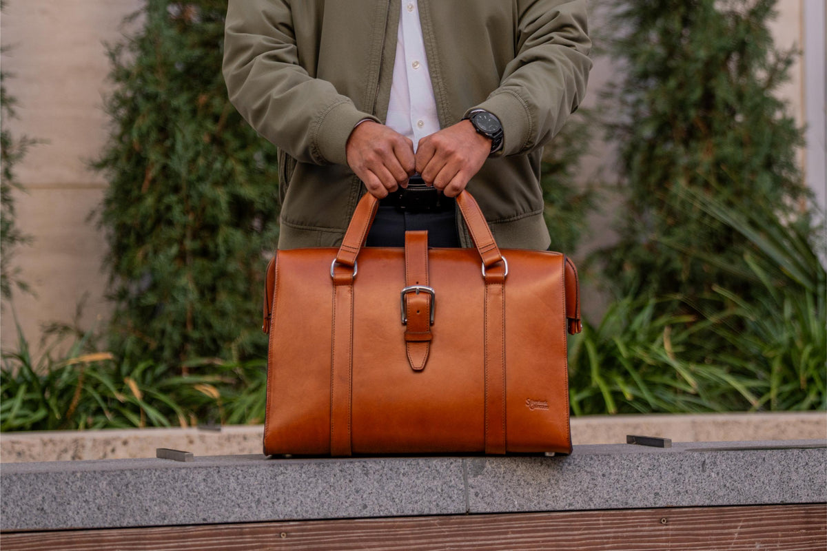 Taylor Duffle Bag in English Tan, held by the handles on a stone ledge outdoors