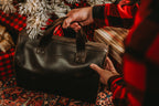 Simple Overnight Leather Duffle Bag in Dark Coffee Brown, held by its handles beside a Christmas tree and wrapped gifts