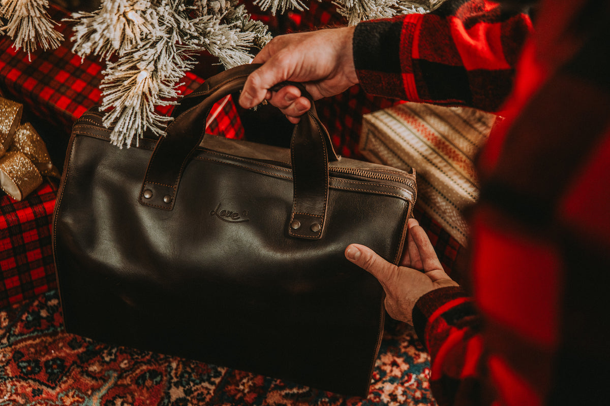 Simple Overnight Leather Duffle Bag in Dark Coffee Brown, held by its handles beside a Christmas tree and wrapped gifts
