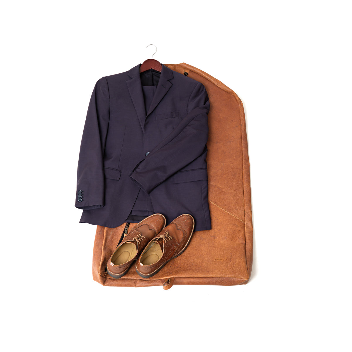 Garment Bag in Tobacco laid flat under a navy suit on a hanger and brown dress shoes