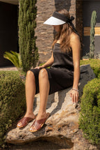 Martha Sandals in Chestnut worn by a woman seated on a garden rock in a black outfit and visor