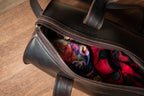 Simple Overnight Leather Duffle Bag in Dark Coffee Brown, partly unzipped with colorful clothing inside on a wood floor