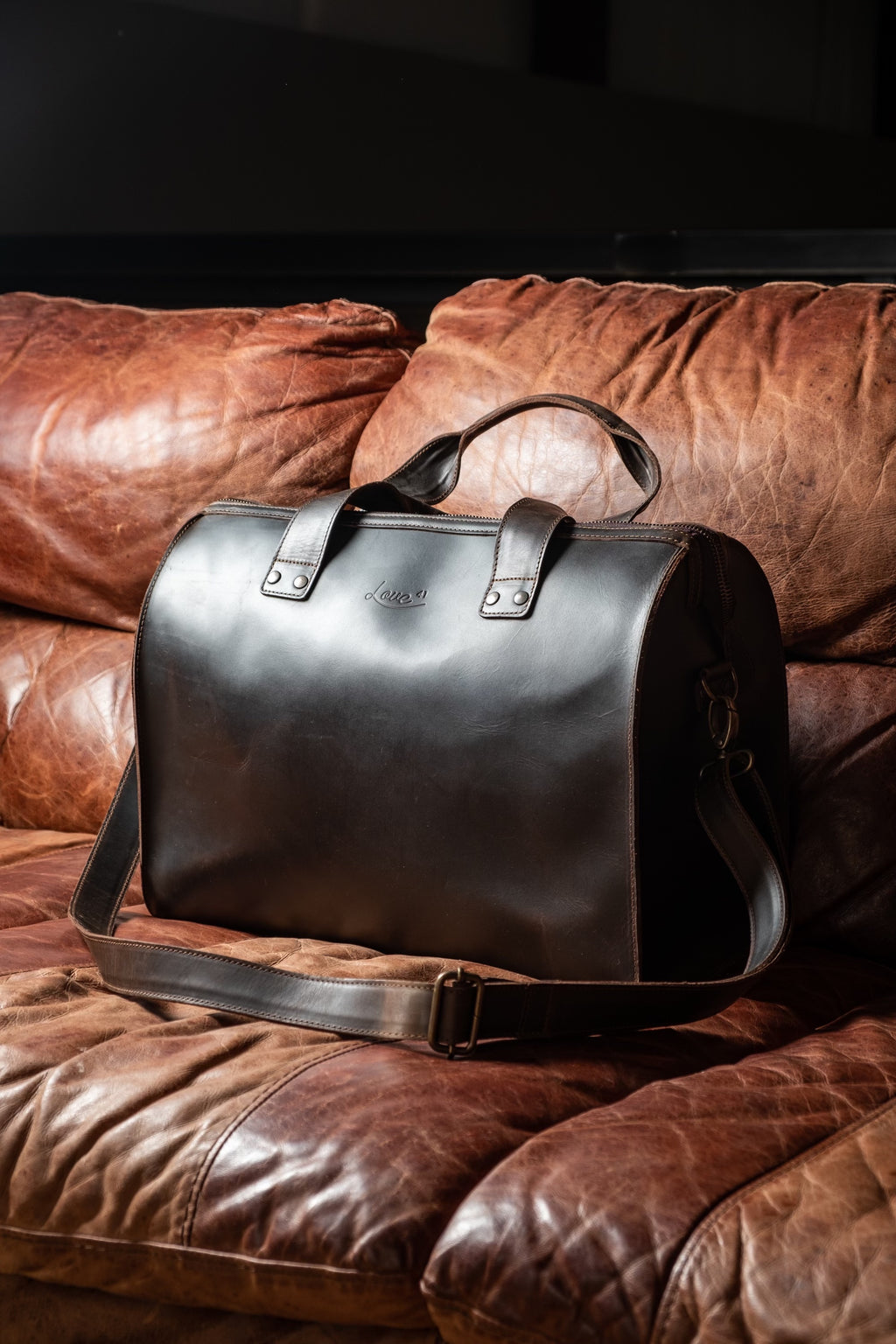 Simple Overnight Leather Duffle Bag in Dark Coffee Brown, resting on a worn brown leather sofa