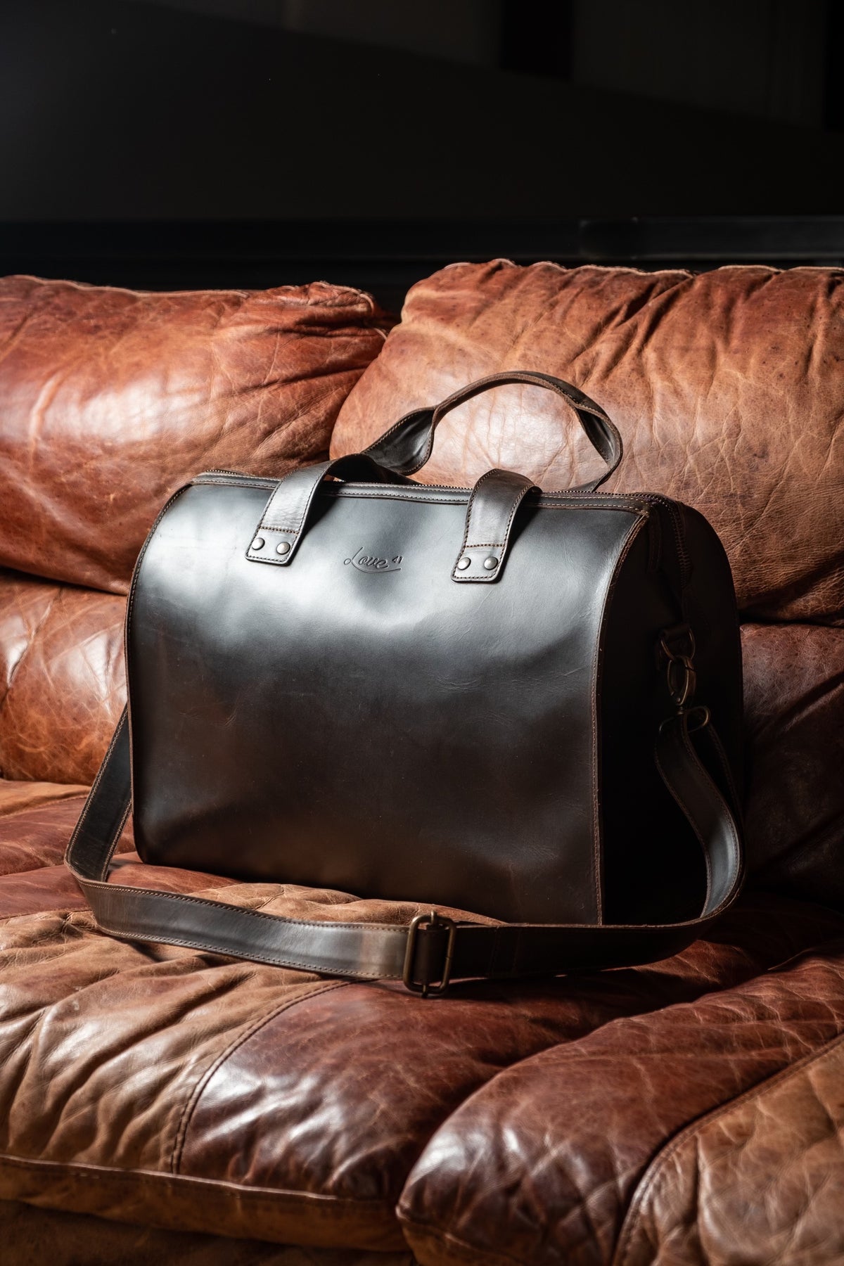 Simple Overnight Leather Duffle Bag in Dark Coffee Brown, resting on a worn brown leather sofa