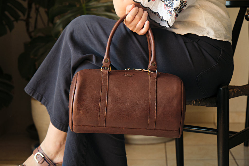 Everyday Barrel Bag in Chestnut, held by its top handles beside a seated woman's knee