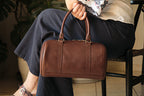 Everyday Barrel Bag in Chestnut, held by its top handles beside a seated woman's knee