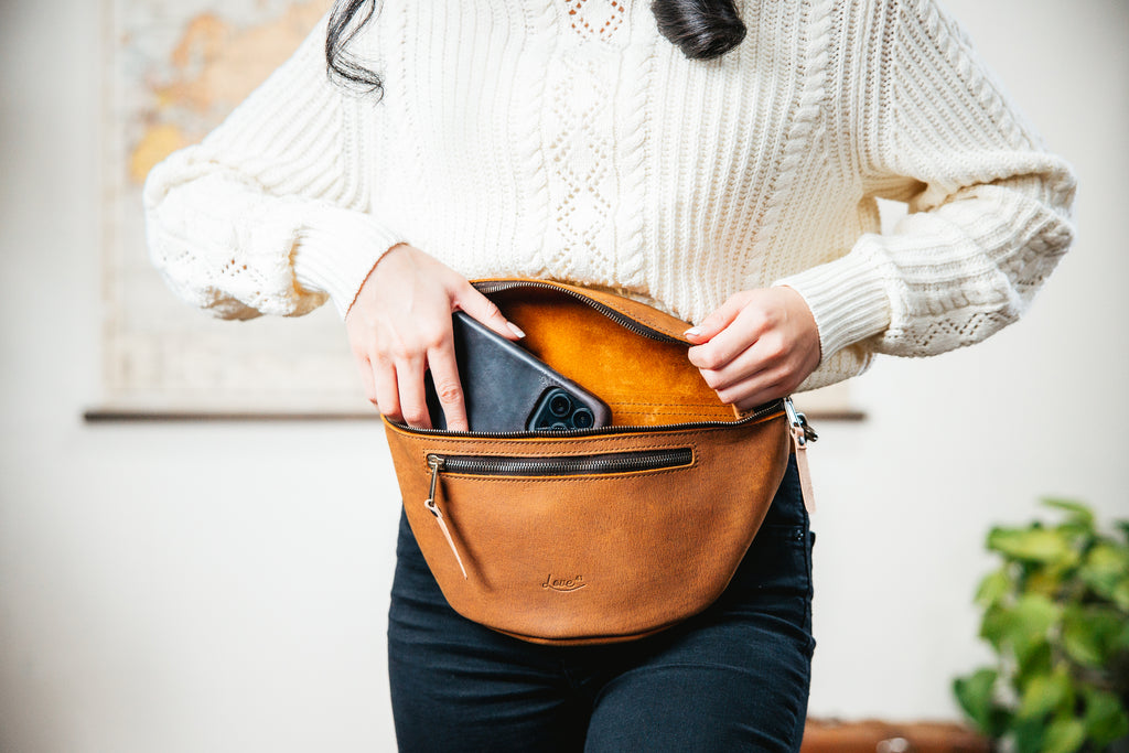 Leather Tumbled Fanny Bag in Tobacco, unzipped at the waist as a woman slides a phone inside