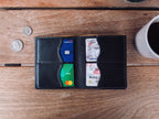 Large Bifold Wallet in Raven Black lying open with cards in the slots, beside coins and a coffee mug