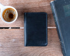 Wycliffe Passport Wallet in Raven Black, closed with debossed logo, between a coffee cup and an old book
