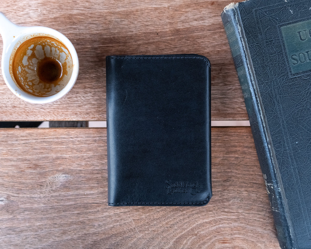 Wycliffe Passport Wallet in Raven Black, closed with debossed logo, between a coffee cup and an old book