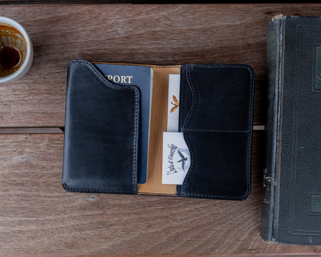 Wycliffe Passport Wallet in Raven Black lying open with a passport and cards, beside a coffee cup and old book