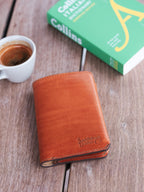 Wycliffe Passport Wallet in English Tan, closed angled view with embossed logo, near a coffee cup and green book