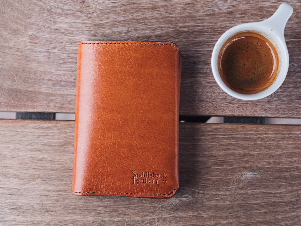 Wycliffe Passport Wallet in English Tan, closed with embossed logo, beside an espresso cup on a wooden table
