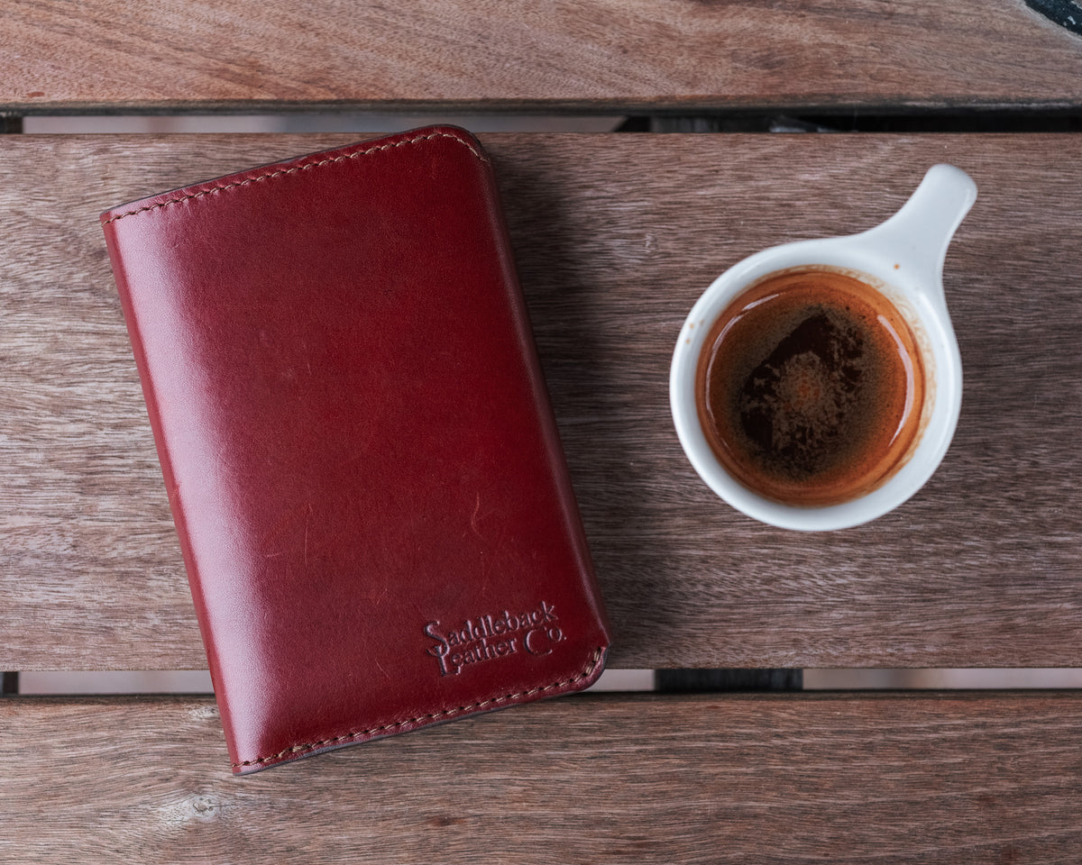 Wycliffe Passport Wallet in Oxblood, closed with embossed logo, beside an espresso cup on a wooden table