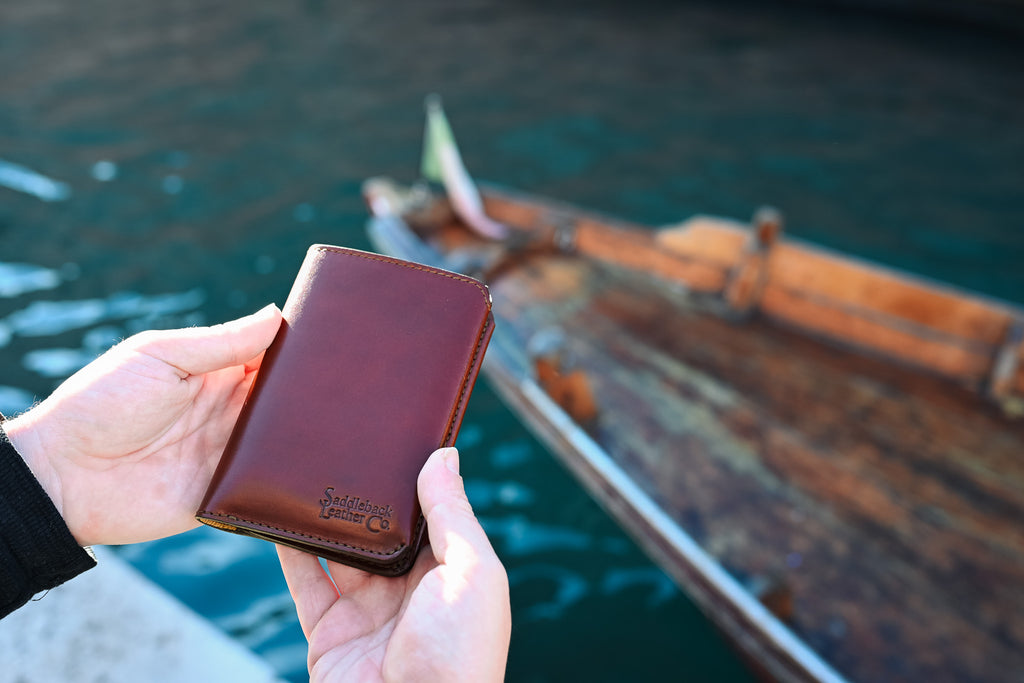 Wycliffe Passport Wallet in Oxblood, closed and held in two hands above the water beside a wooden boat