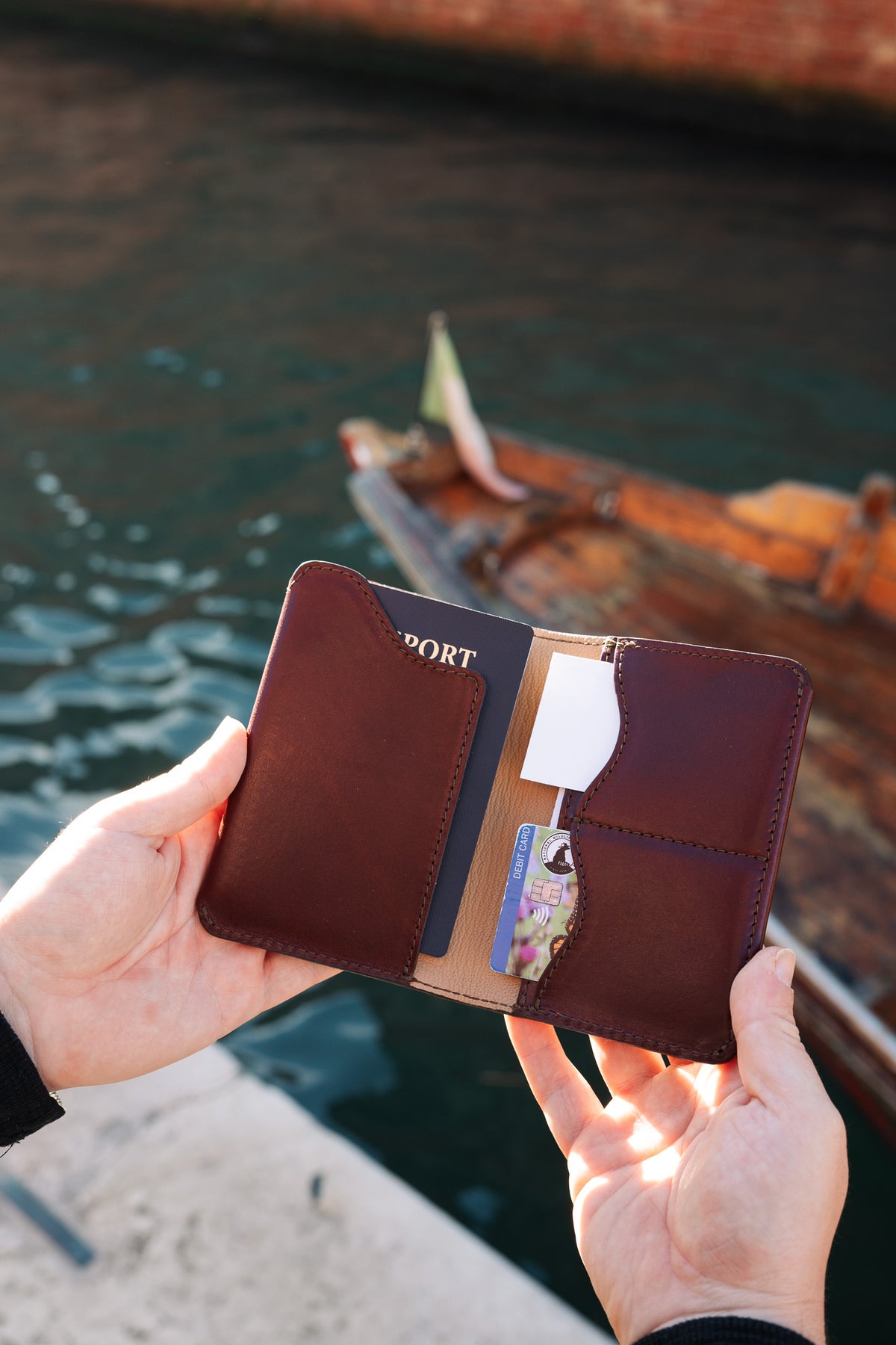 Wycliffe Passport Wallet in Oxblood held open in sunlight with a passport and cards, a wooden boat behind
