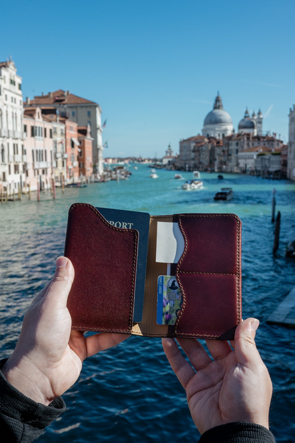 Wycliffe Passport Wallet in Oxblood held open in front of a canal lined with historic buildings and a domed church