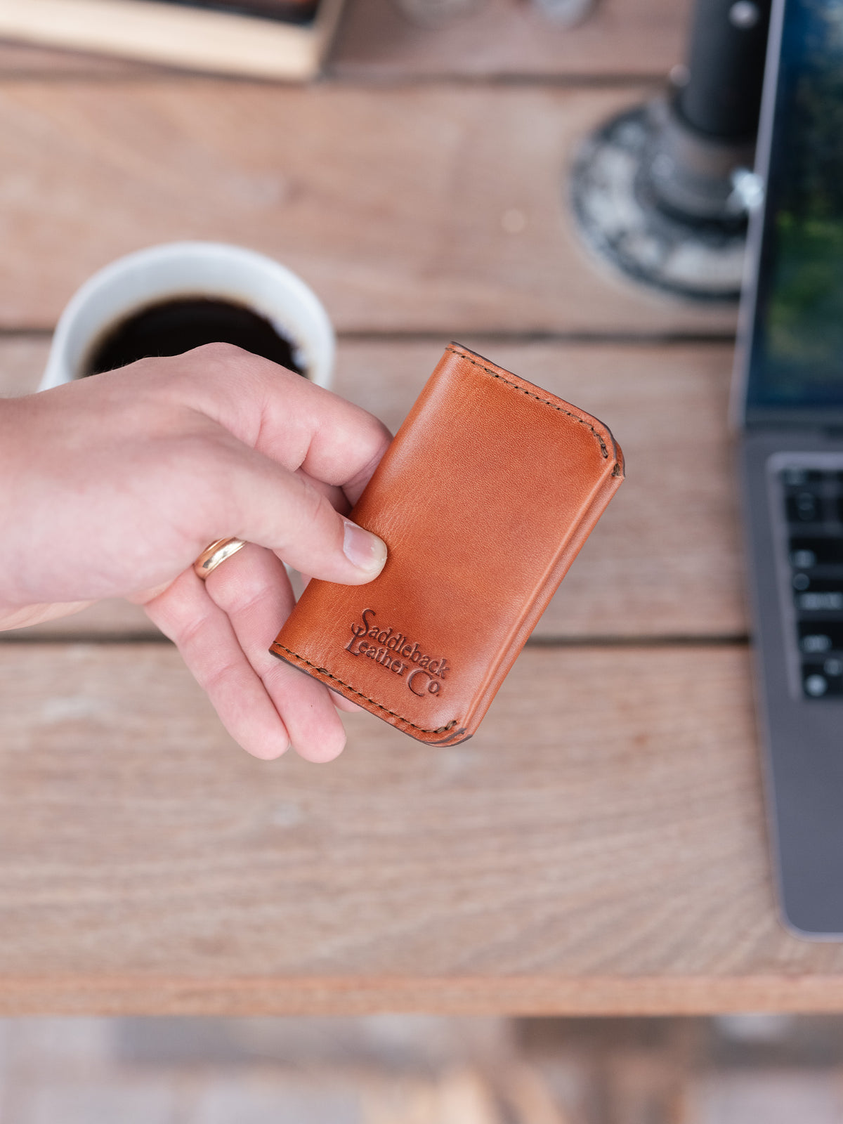 Slim Business Card Holder in English Tan, held in hand above a desk with coffee and a laptop