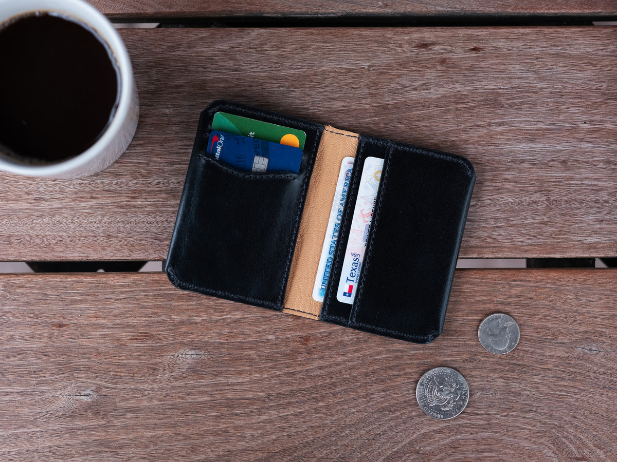 Slim Bifold Wallet in Black, open with cards on a wooden table beside coffee and coins