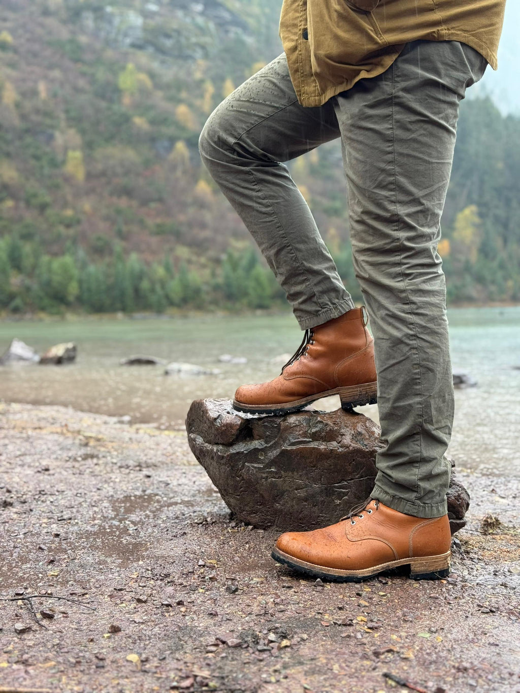 Huntsman Boots in Natural, one foot on a rock by a lake with forested hills behind