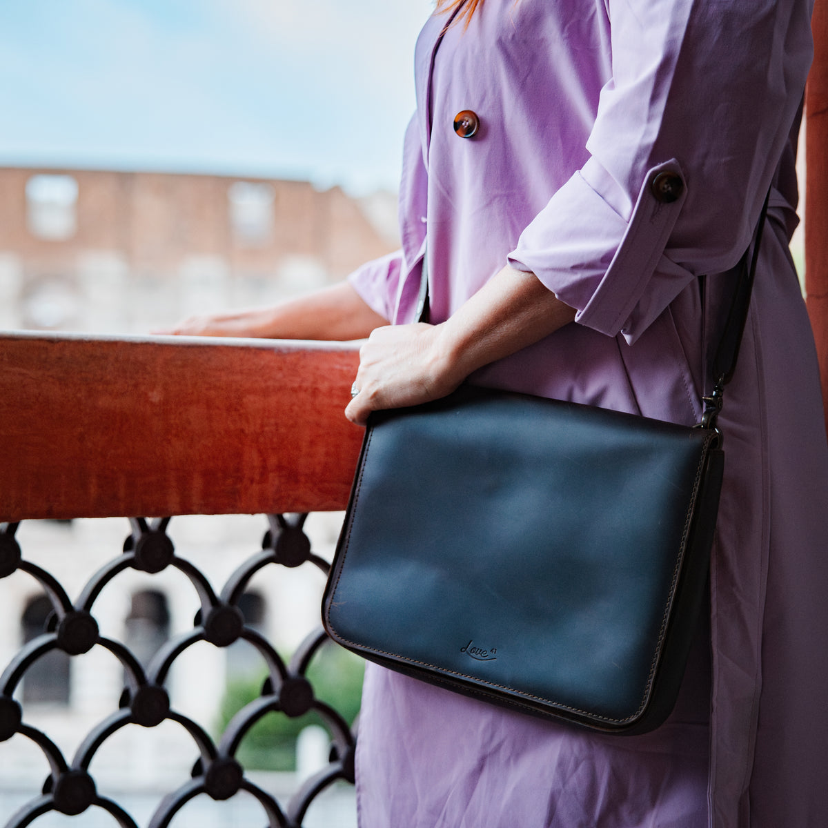 Magnetic Fold Over Bag Large in a dark finish, carried on the shoulder by a woman in a lilac coat on a balcony