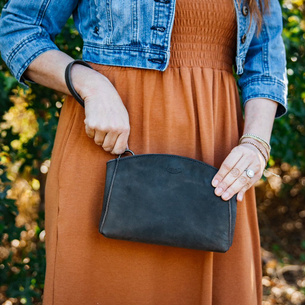 Leather Organizer Clutch in charcoal gray, held by a woman in a rust dress and denim jacket with wrist strap on her arm