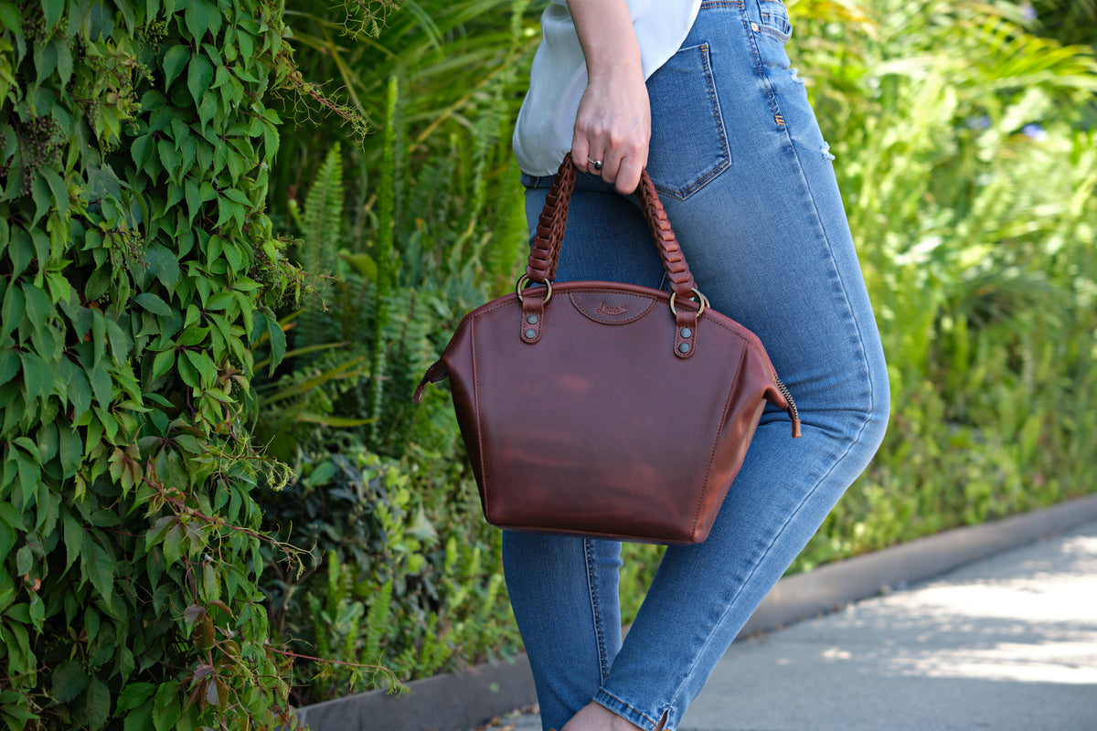 Dovetail Satchel in Chestnut, carried by its braided handles along a leafy garden path