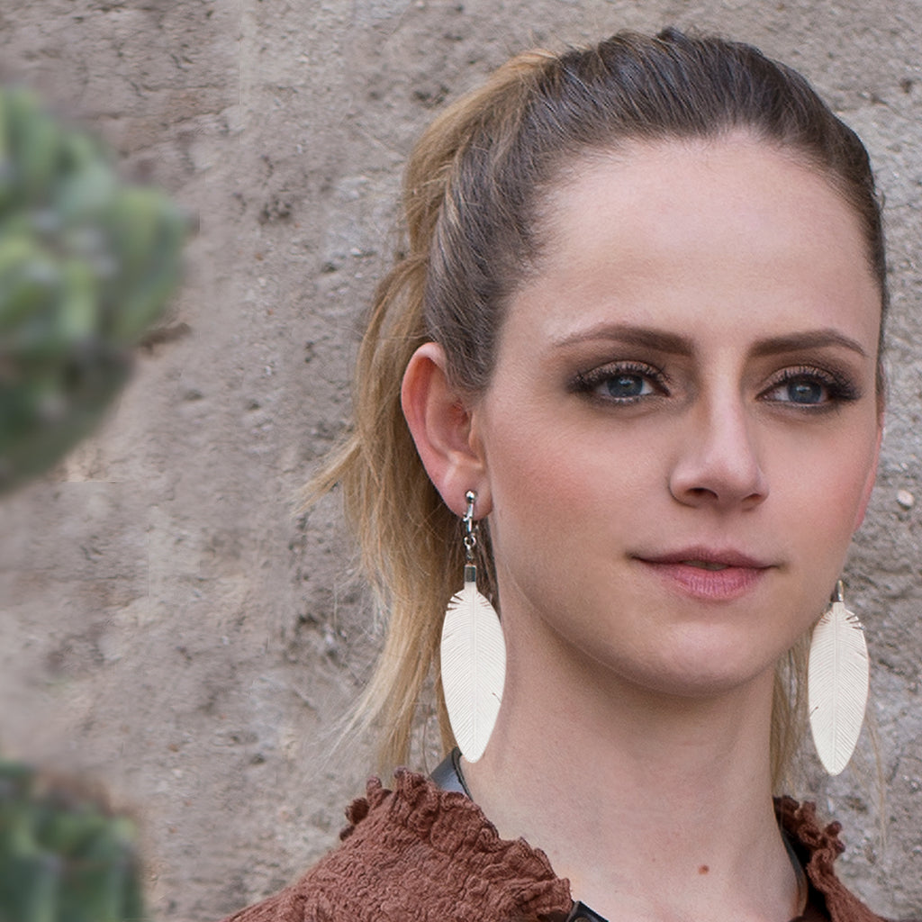Leather Feather Earrings in White worn by a woman with her hair in a ponytail against a stone wall