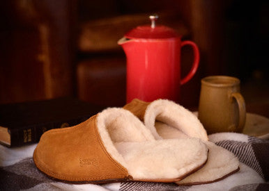 Men's Sheepskin Slippers in Tobacco on a table beside a red teapot and mug