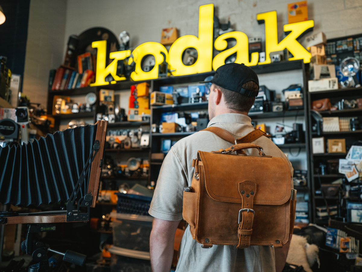 Photographer's Satchel in Tobacco, worn as a backpack by a man inside a vintage camera shop