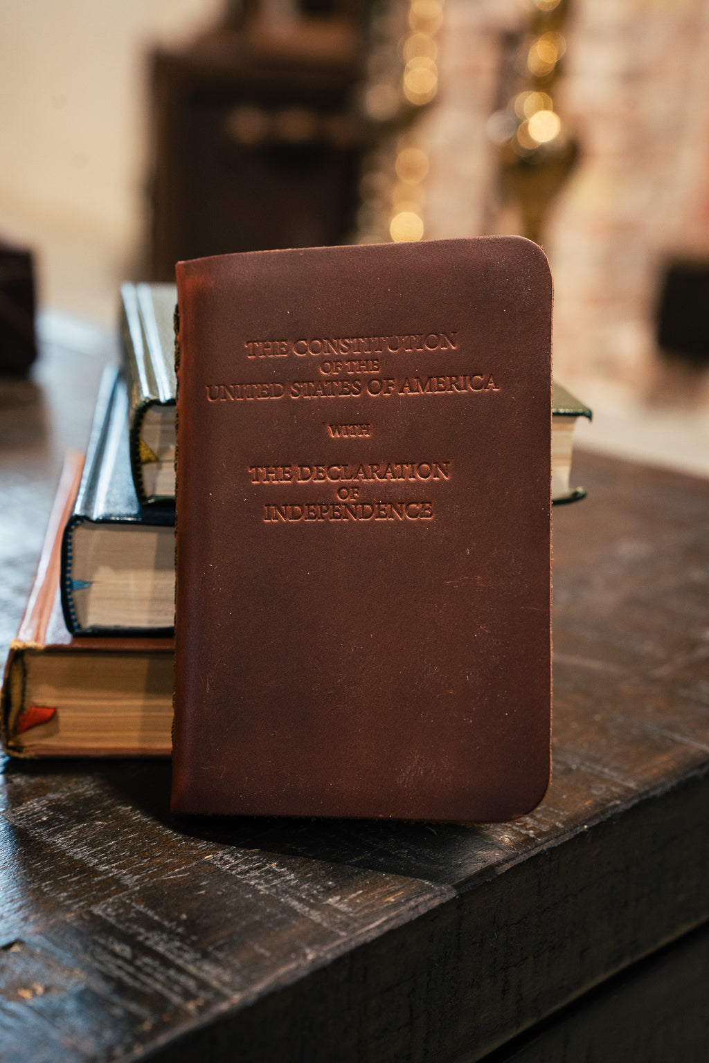Pocket Declaration/Constitution in Dark Coffee Brown standing against a stack of books on a wooden table