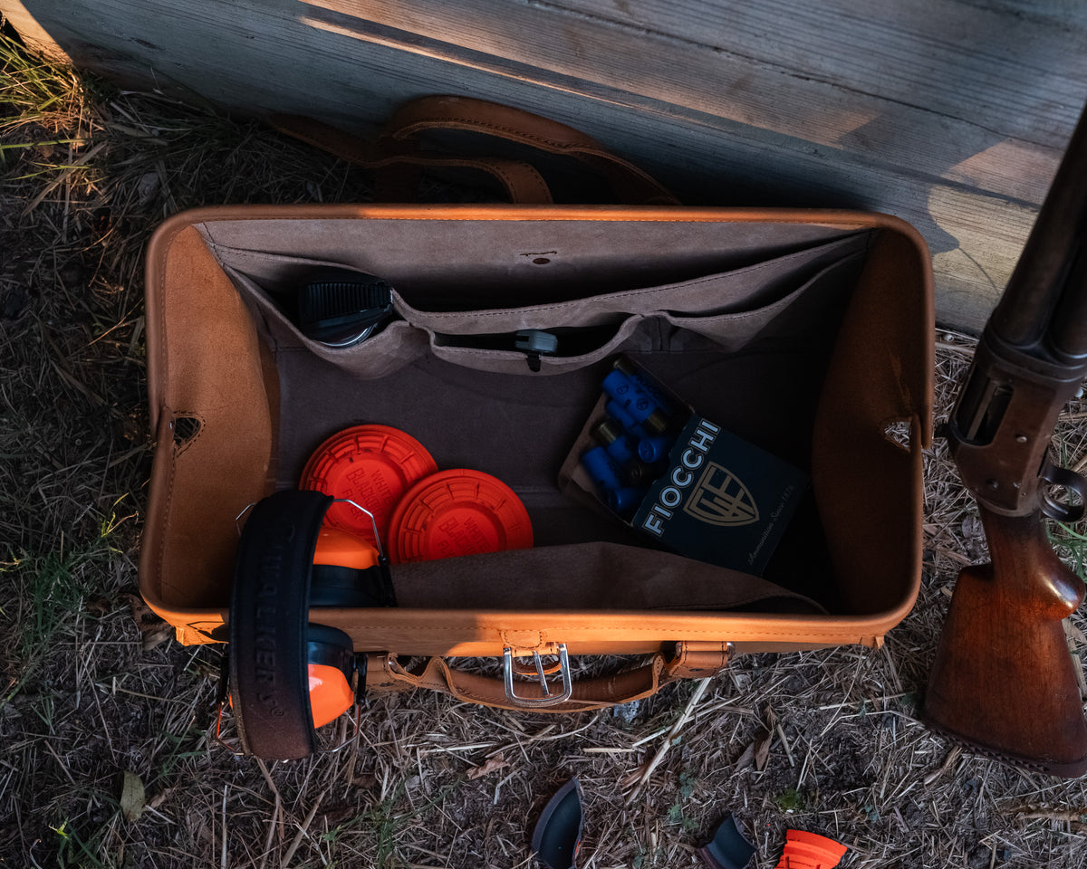 Leather Range and Tool Bag in Tobacco, open on grass with clay targets, ear muffs and shells beside a shotgun