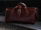 Leather Range and Tool Bag in Chestnut, front view on a dark wooden tailgate with top handles and buckle strap