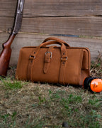 Leather Range and Tool Bag in Tobacco on grass against a wooden wall beside a shotgun and orange ear muffs