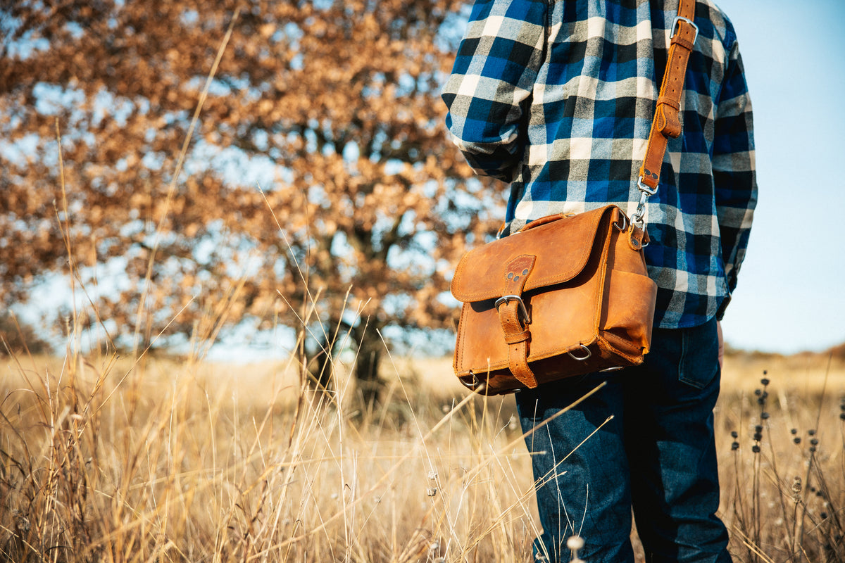 Photographer's Satchel in Tobacco, worn crossbody by a man in a plaid shirt in a dry grass field