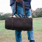 A man holding the Range and Tool Tote in Dark Coffee Brown by its handles, packed with tools, in a field