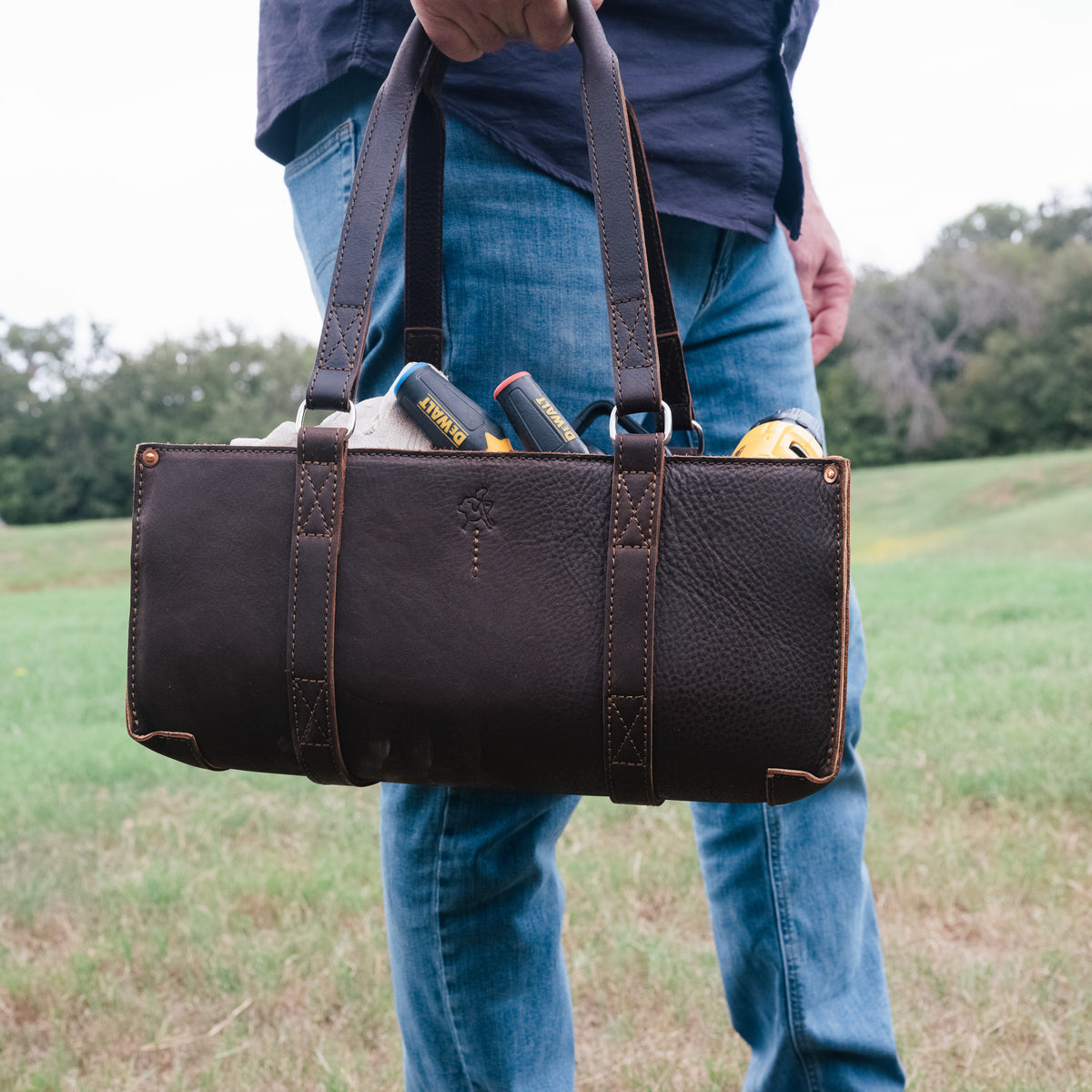 A man holding the Range and Tool Tote in Dark Coffee Brown by its handles, packed with tools, in a field